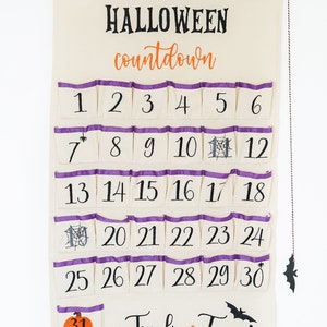 May include: A Halloween countdown calendar made of cream-colored fabric with purple trim on the pockets. The calendar has 31 numbered pockets, with the words "HALLOWEEN countdown" at the top and "Trick or Treat" at the bottom. A bat decoration hangs from the side.