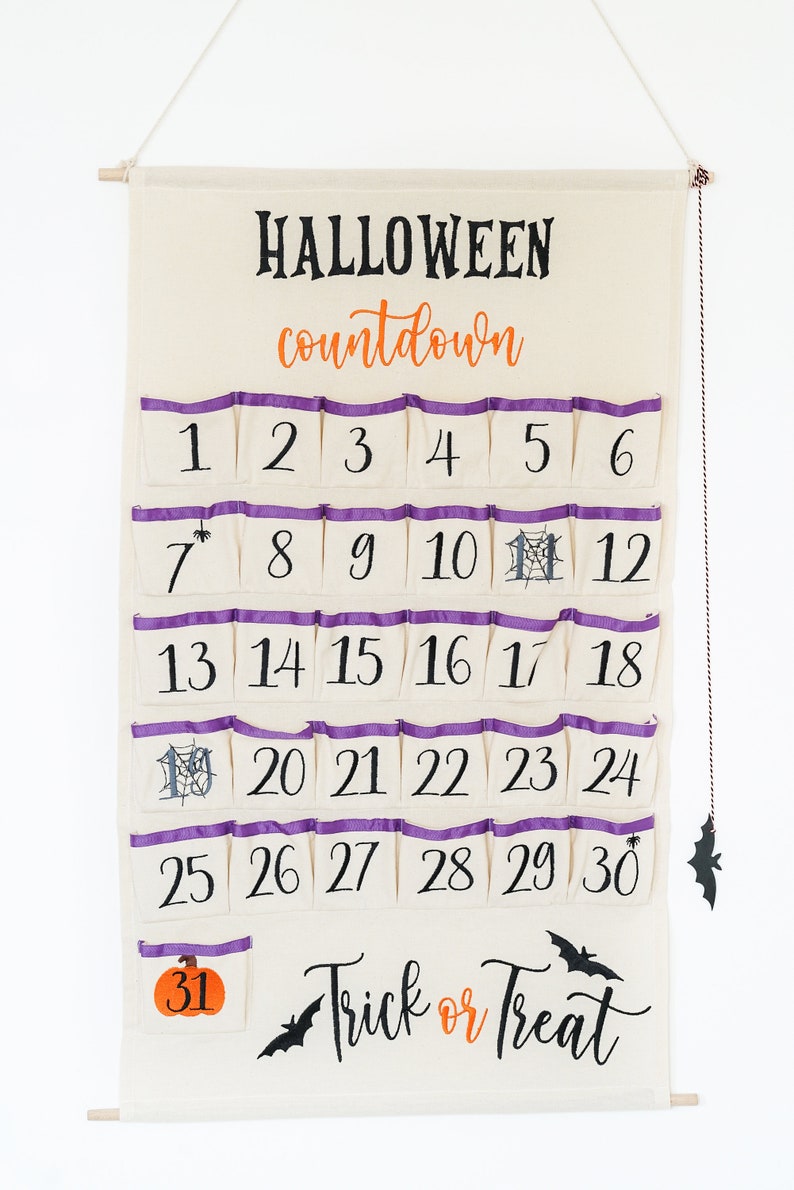 May include: A Halloween countdown calendar made of cream-colored fabric with purple trim on the pockets. The calendar has 31 numbered pockets, with the words "HALLOWEEN countdown" at the top and "Trick or Treat" at the bottom. A bat decoration hangs from the side.
