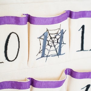 May include: Close-up of a cream-colored fabric advent calendar with purple ribbon trim. The calendar has pockets with embroidered numbers 10, 11, and 12 in black. Number 11 features a gray spiderweb design.