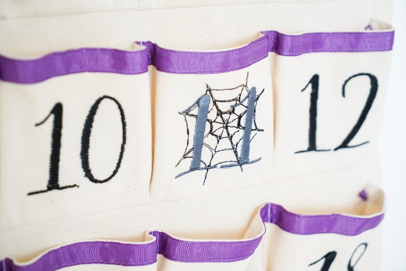May include: Close-up of a cream-colored fabric advent calendar with purple ribbon trim. The calendar has pockets with embroidered numbers 10, 11, and 12 in black. Number 11 features a gray spiderweb design.