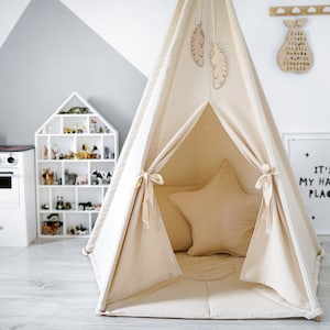 Könnte beinhalten: Ein beigefarbenes Tipi-Zelt aus Segeltuch für Kinder mit einem sternförmigen Kissen im Inneren. Das Tipi hat hölzerne Federn, die von oben hängen, und ist in einem Raum mit einer weißen Spielküche und einem weißen Puppenhaus aufgestellt.