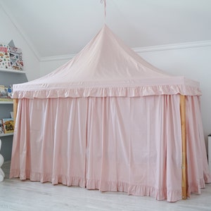 May include: A light pink fabric bed canopy with a pointed top and ruffled trim edges. The canopy is draped over a wooden frame structure, creating a tent-like enclosure with long, flowing curtains that reach the floor on all visible sides.