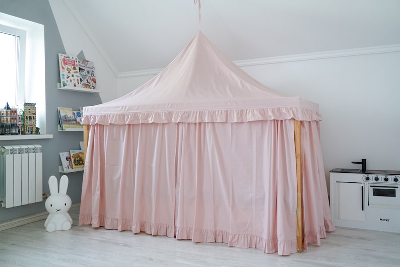 May include: A light pink fabric bed canopy with a pointed top and ruffled trim edges. The canopy is draped over a wooden frame structure, creating a tent-like enclosure with long, flowing curtains that reach the floor on all visible sides.