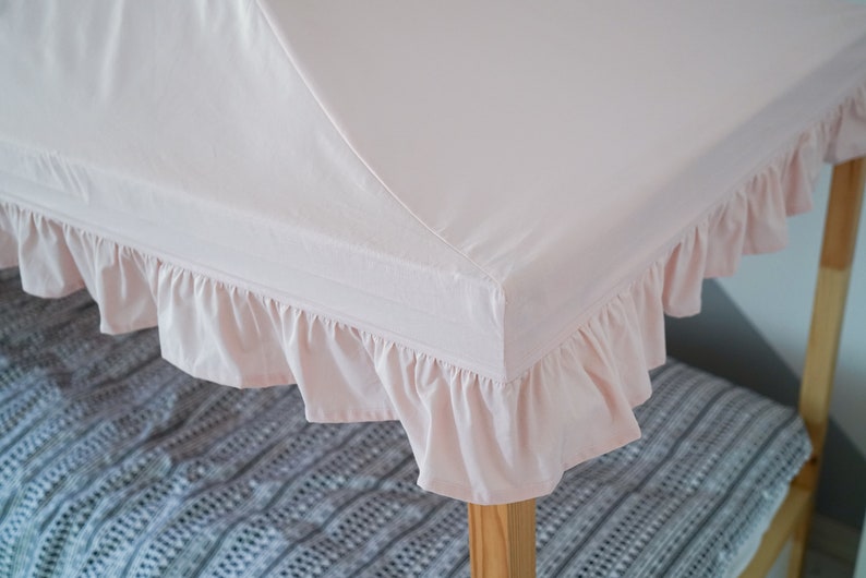 May include: A light pink fabric canopy cover featuring a ruffled edge trim. The canopy is designed to fit over a wooden frame structure, providing a soft, solid-colored textile top with gathered fabric detailing along the perimeter of the frame.