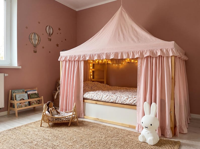 May include: A light pink fabric bed canopy with a ruffled trim draped over a wooden bed frame. The canopy features a pointed top and side curtains. Inside the bed, a patterned mattress is visible, illuminated by a string of warm white fairy lights.