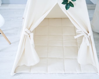 May include: A white canvas teepee tent with a white quilted floor mat. The teepee has two flaps that are tied together with white strings. The teepee is set up on a white wooden floor.