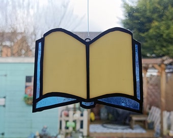 Op de afbeelding: Een gele glas-in-lood boek ornament met een blauwe rand, hangend voor een raam.