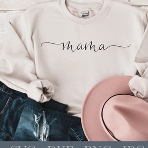 Puede incluir: Una sudadera blanca con la palabra "mama" escrita en cursiva en la parte delantera. La sudadera está doblada y colocada sobre una superficie de madera con un par de pantalones vaqueros azules y un sombrero rosa.