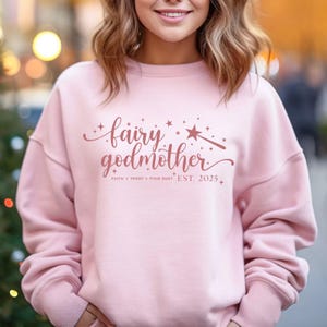 May include: A pale pink sweatshirt featuring the text "fairy godmother" in a script font, with the words "faith, trust, pixie dust, est. 2025". The design includes stars and a wand.