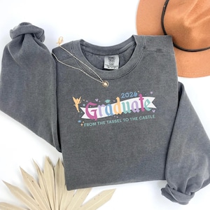 Peut inclure: Un sweat-shirt gris foncé avec le mot "Graduate" en texte coloré, l'année "2026" et la phrase "From the Tassel to the Castle". Un collier doré et un chapeau marron sont également visibles.