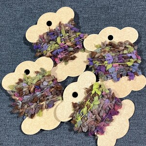 May include: Five skeins of colorful yarn are displayed on cloud-shaped cardboard tags. The yarn is a mix of purple, green, brown, and silver, with a textured, fluffy appearance. The tags have a small hole at the top for hanging.