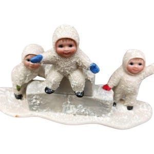 May include: Three small, white porcelain figurines of children wearing winter clothing and playing in the snow. The children are wearing blue and red mittens and are standing on a snow-covered base.