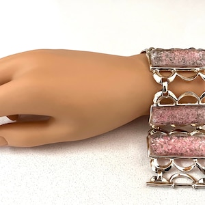 May include: A silver-toned bracelet with three rectangular pink and gray stone panels. The panels are connected by decorative silver links. The bracelet is displayed on a mannequin hand.