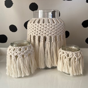 May include: Three glass candle holders with crocheted covers and fringe. The covers are cream coloured and the fringe is long and flowing. The candle holders are on a white surface with black polka dots.
