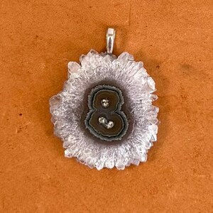 May include: A unique pendant featuring a natural, layered stone with a central, dark-colored design. The outer edges are a light, crystalline color, and the pendant has a silver-colored bail for attaching to a chain. The stone has a natural, organic shape.