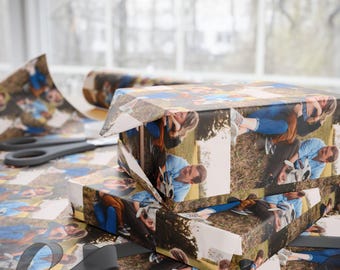 May include: A wrapped gift box with custom photo wrapping paper. The paper features multiple images of a couple. Scissors and a roll of wrapping paper are on the table. A grey ribbon is wrapped around the gift.