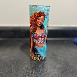 Puede incluir: Un vaso de color turquesa y azul con una ilustraci&oacute;n colorida de una sirena con cabello rojo y un sujetador de concha rosa. El texto "La Sirenita" est&aacute; impreso en la parte inferior del vaso.