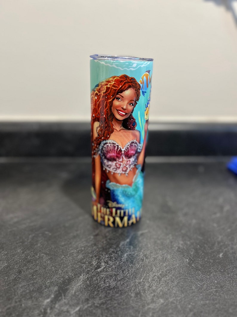 Puede incluir: Un vaso de color turquesa y azul con una ilustraci&oacute;n colorida de una sirena con cabello rojo y un sujetador de concha rosa. El texto "La Sirenita" est&aacute; impreso en la parte inferior del vaso.