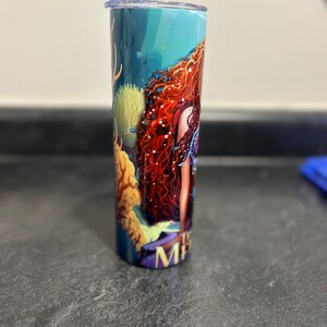 Puede incluir: Un vaso de acero inoxidable con un dise&ntilde;o de sirena. El vaso presenta un fondo azul, verde y amarillo con una sirena de pelo rojo con un collar de perlas. El texto "Mermaid" es visible en la parte inferior del vaso.