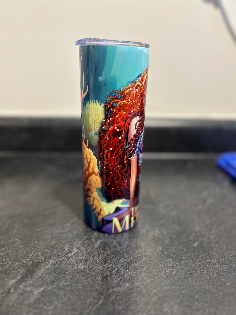 Puede incluir: Un vaso de acero inoxidable con un dise&ntilde;o de sirena. El vaso presenta un fondo azul, verde y amarillo con una sirena de pelo rojo con un collar de perlas. El texto "Mermaid" es visible en la parte inferior del vaso.