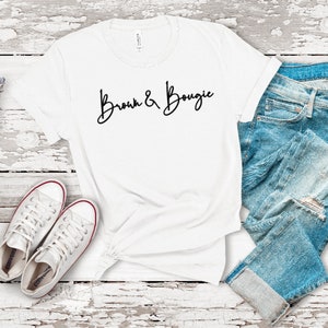 May include: White t-shirt with the text "Bronx & Bougie" in black script lettering. The shirt is laid flat on a white wooden surface with a pair of white sneakers and blue jeans.