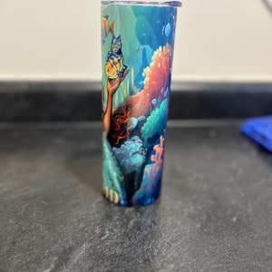 Puede incluir: Un vaso azul y verde con un dise&ntilde;o de sirena y peces. La sirena sostiene un pez amarillo en su mano. El vaso tiene una tapa transparente.