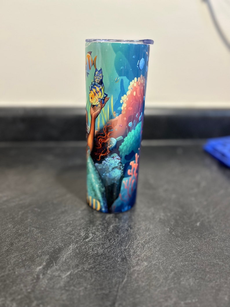 Puede incluir: Un vaso azul y verde con un dise&ntilde;o de sirena y peces. La sirena sostiene un pez amarillo en su mano. El vaso tiene una tapa transparente.