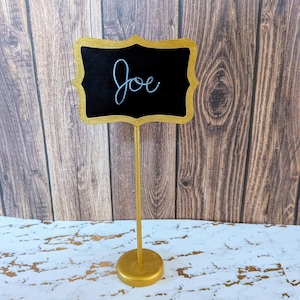May include: A gold-colored place card holder with a black chalkboard-style sign. The sign has a gold ornate border and the name "Joe" written in cursive. The base and stand are also gold.