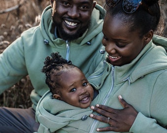 May include: A family wearing matching green hooded sweatshirts. The parents are sitting on the ground, and the child is being held in the mother's arms. The child is wearing a green hooded sweatshirt with a zipper and buttons.