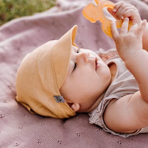 Könnte beinhalten: Ein Baby trägt eine hellbraune Mütze aus Leinen mit einem kleinen Herzlogo an der Seite, liegt auf einer rosa gestrickten Decke und hält einen gelben Kauspielring.