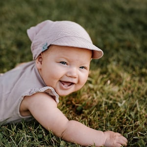 May include: A baby wearing a light pink hat with a light blue logo on the side, is lying on its tummy on green grass. The baby is smiling and looking at the camera.