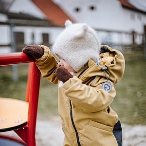 Peut inclure: Un jeune enfant portant une combinaison de neige marron et jaune avec un bonnet en laine blanc et des mitaines marron. L'enfant se tient sur une aire de jeux près d'un jouet tournant rouge et bleu.