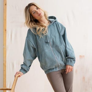 Könnte beinhalten: Hellblaues Cord-Sweatshirt mit Reißverschluss, Kapuze mit Kordelzug und passender Hose. Das Model trägt braune Schuhe und lehnt sich an einen Korbstuhl. Das Outfit ist lässig und bequem.