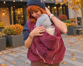 May include: A deep burgundy corduroy babywearing jacket featuring a front pouch for an infant, a hood, and a zipper closure. The jacket is worn over a dark blue long-sleeve shirt, paired with brown trousers and a dark blue ribbed knit beanie.