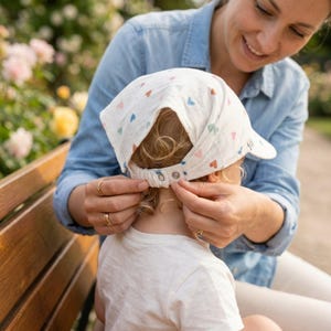 Op de afbeelding: Een witte baby zonnehoed met een klep en een reeks kleine, kleurrijke hartjes. De hoed wordt vastgemaakt door een persoon die een spijkerblouse draagt. De achtergrond toont een park met bloemen.