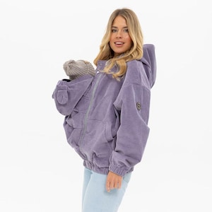 Puede incluir: Una chaqueta de pana lavanda con capucha y cremallera frontal. La chaqueta tiene un corte holgado, bolsillos delanteros y un pequeño emblema en la manga. Un gorro gris es visible en el bolsillo de la chaqueta. La modelo lleva vaqueros azul claro.