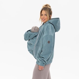 Puede incluir: Chaqueta de pana color turquesa con capucha y portabebés incorporado. La chaqueta tiene cierre de cremallera y un pequeño parche en el pecho. Un bebé está acurrucado dentro del portabebés, con un gorro gris.