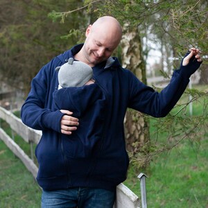 Puede incluir: Un hombre lleva una sudadera con capucha azul oscuro con un portabeb&eacute;s incorporado. La sudadera tiene una cremallera y una capucha. El hombre lleva un beb&eacute; en el portabeb&eacute;s. El beb&eacute; lleva una gorra gris.