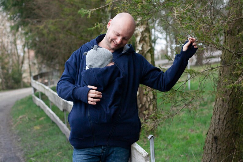 Puede incluir: Un hombre lleva una sudadera con capucha azul oscuro con un portabeb&eacute;s incorporado. La sudadera tiene una cremallera y una capucha. El hombre lleva un beb&eacute; en el portabeb&eacute;s. El beb&eacute; lleva una gorra gris.