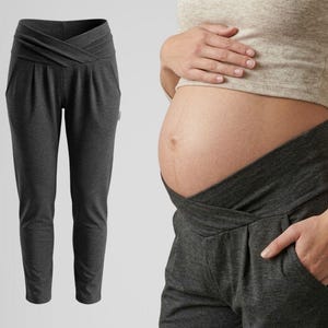 May include: Dark gray maternity pants with a crossover waistband and pockets. The pants are shown on a white background and also being worn. The design features a comfortable, stretchy fabric suitable for pregnancy. The pants are designed to accommodate a growing belly.