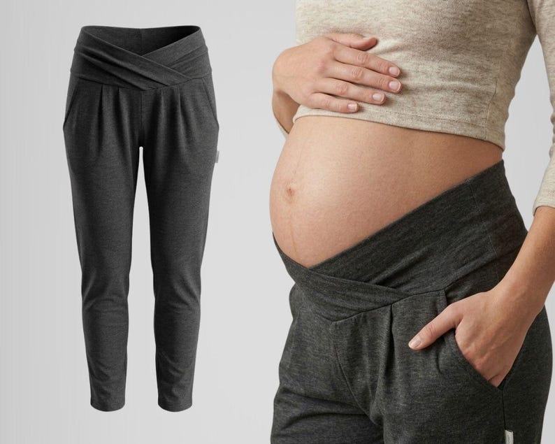 May include: Dark gray maternity pants with a crossover waistband and pockets. The pants are shown on a white background and also being worn. The design features a comfortable, stretchy fabric suitable for pregnancy. The pants are designed to accommodate a growing belly.