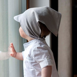 May include: A young child wearing a grey bandana and a white shirt looks out of a window.