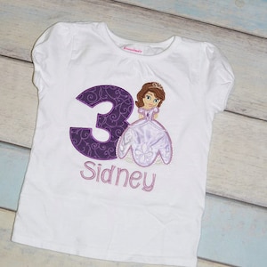 May include: White t-shirt with a purple number 3 and a purple embroidered image of a princess in a lavender dress. The name "Sidney" is embroidered below the number.