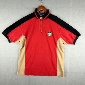 May include: A short-sleeved polo shirt in red, black, and tan. The shirt has a zip-up collar, a small embroidered emblem, and color-blocked panels. The sleeves are black, and the sides are tan.