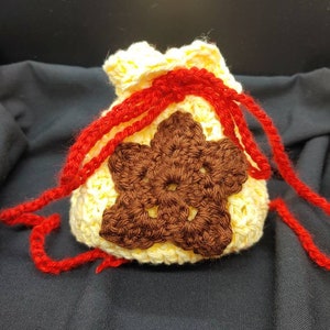 May include: A crocheted drawstring pouch in cream with a brown star-shaped applique. Red yarn forms the drawstring and accents the pouch. The pouch is handmade and has a textured appearance.