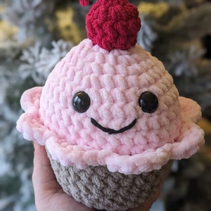 May include: A pink and brown crocheted cupcake plush toy with a red cherry on top. The cupcake has a smiling face with black eyes.