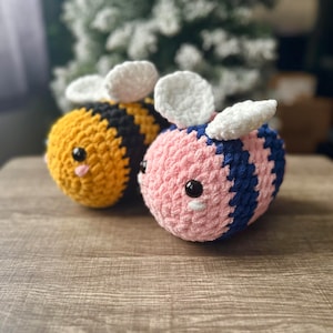 Peut inclure: Deux peluches abeilles en crochet, l'une jaune et noire, l'autre rose et bleue, avec des ailes blanches.