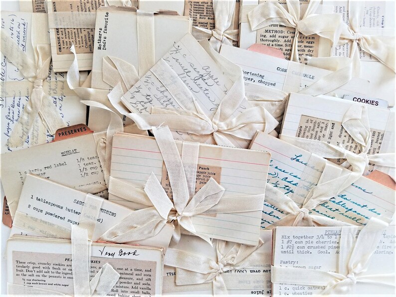 pack of 10 vintage recipe cards tied with ribbon.  The cards are filled with handwritten recipes and feature titles such as 'Mother's Favorite,' 'Cookies,' and 'Preserves.'  The recipes are written in blue or black ink on off-white or light-cream colored recipe cards. Some cards show typed recipes. The overall aesthetic is reminiscent of antique cookbooks and home-style baking.