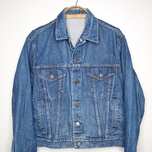 May include: A medium-wash denim jacket with a classic collar, button-up front, and two chest pockets. The jacket has a standard fit and is hanging on a wooden hanger. The denim is a solid blue color.