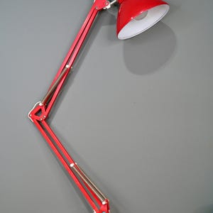 May include: A red desk lamp with a white interior shade. The lamp has a flexible, articulated arm and a red base. The lamp is plugged into a white electrical outlet. The lamp is on a wooden surface.